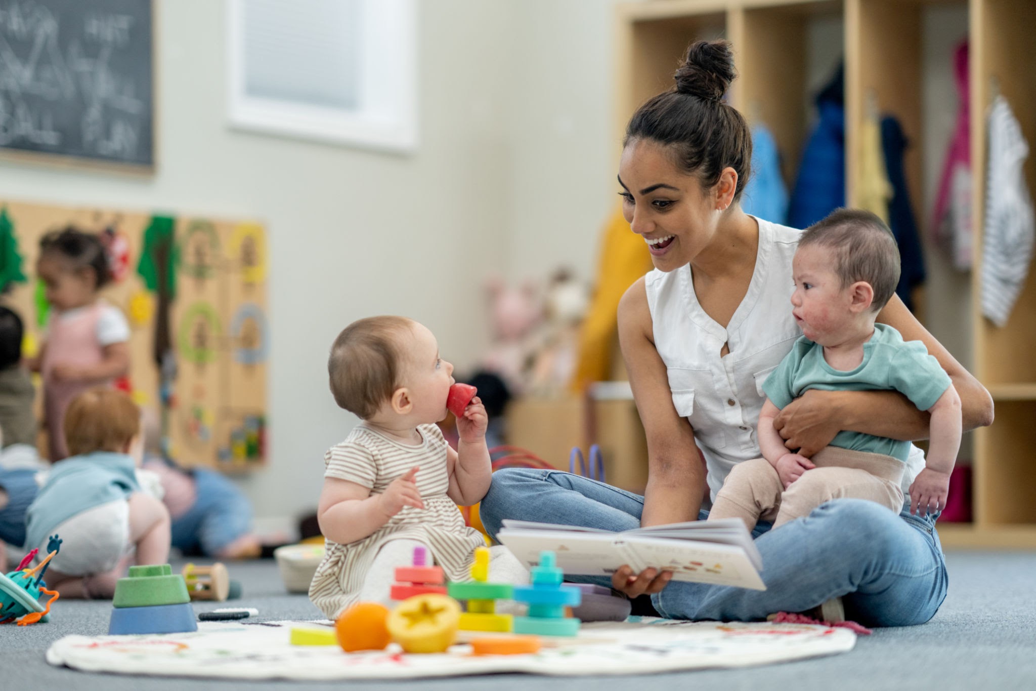 infantsdaycareaz A small group of babies sit on the floor playing with various toys while their teacher supervises and engages them. They are each dressed casually and appear content.