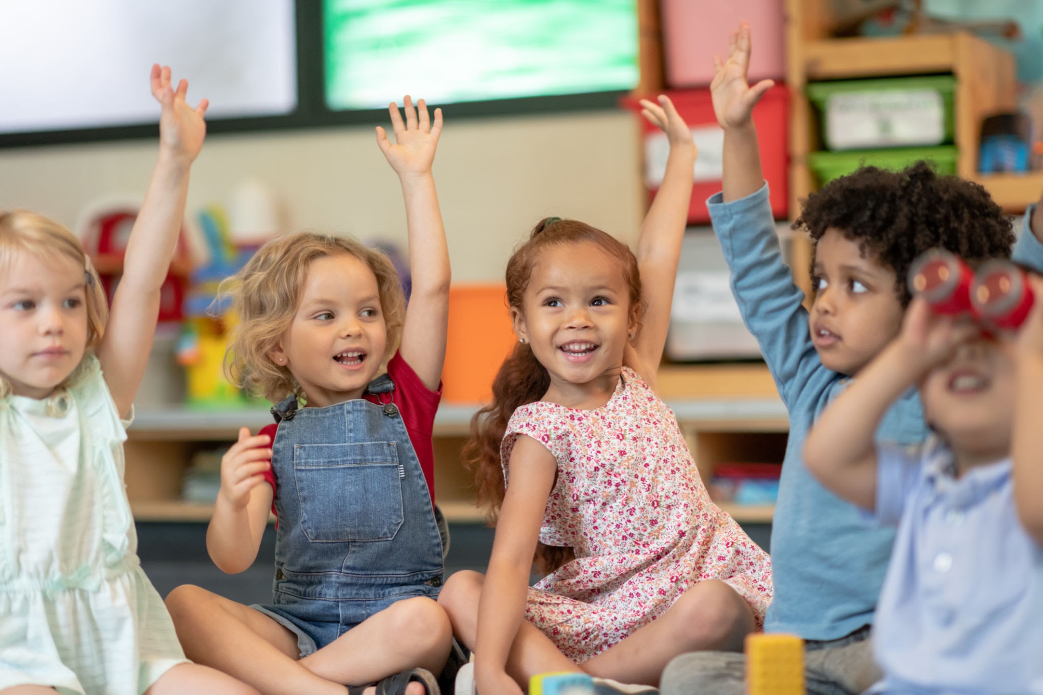 preschooldaycare A multi-ethnic group of preschool students is sitting with their legs crossed on the floor in their classroom. The teacher is sitting on the floor facing the children. The happy kids are smiling and listening to the teacher. They have their arms raised and are eager to answer the teacher's question.